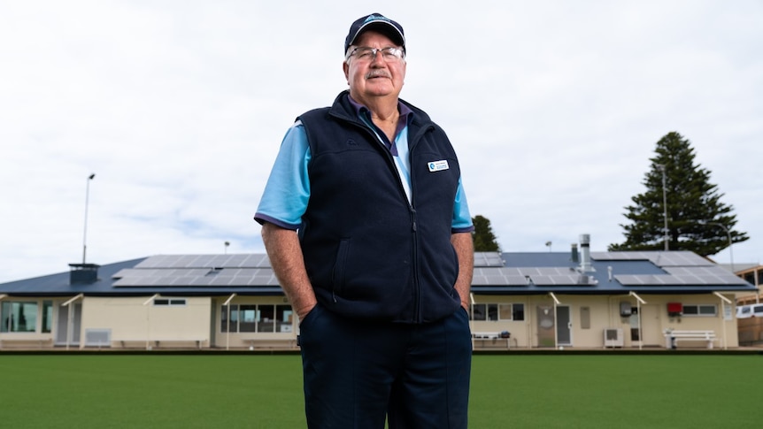 A man wearing a cap, glasses, a blue shirt and a navy vest stands on a bowling green with the clubrooms behind him