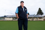 A man wearing a cap, glasses, a blue shirt and a navy vest stands on a bowling green with the clubrooms behind him