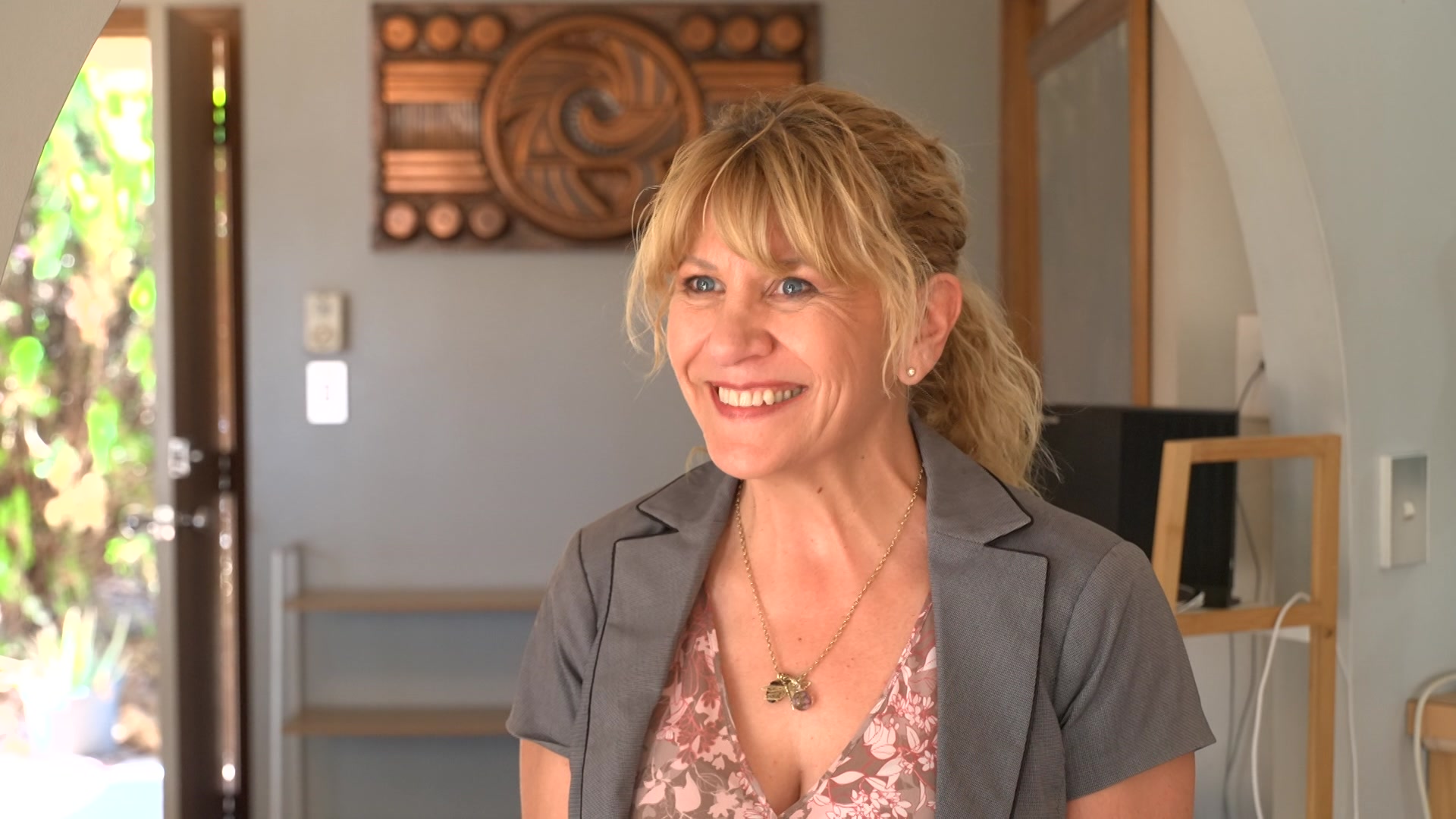 Una mujer sonriente con cabello rubio y vestida con una chaqueta gris de manga corta. Detrás de ella hay obras de arte y muebles.