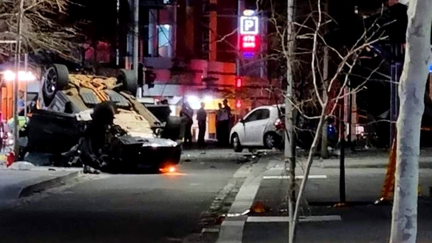 A long-range photo of a car flipped on its roof with a group of emergency workers in the background.