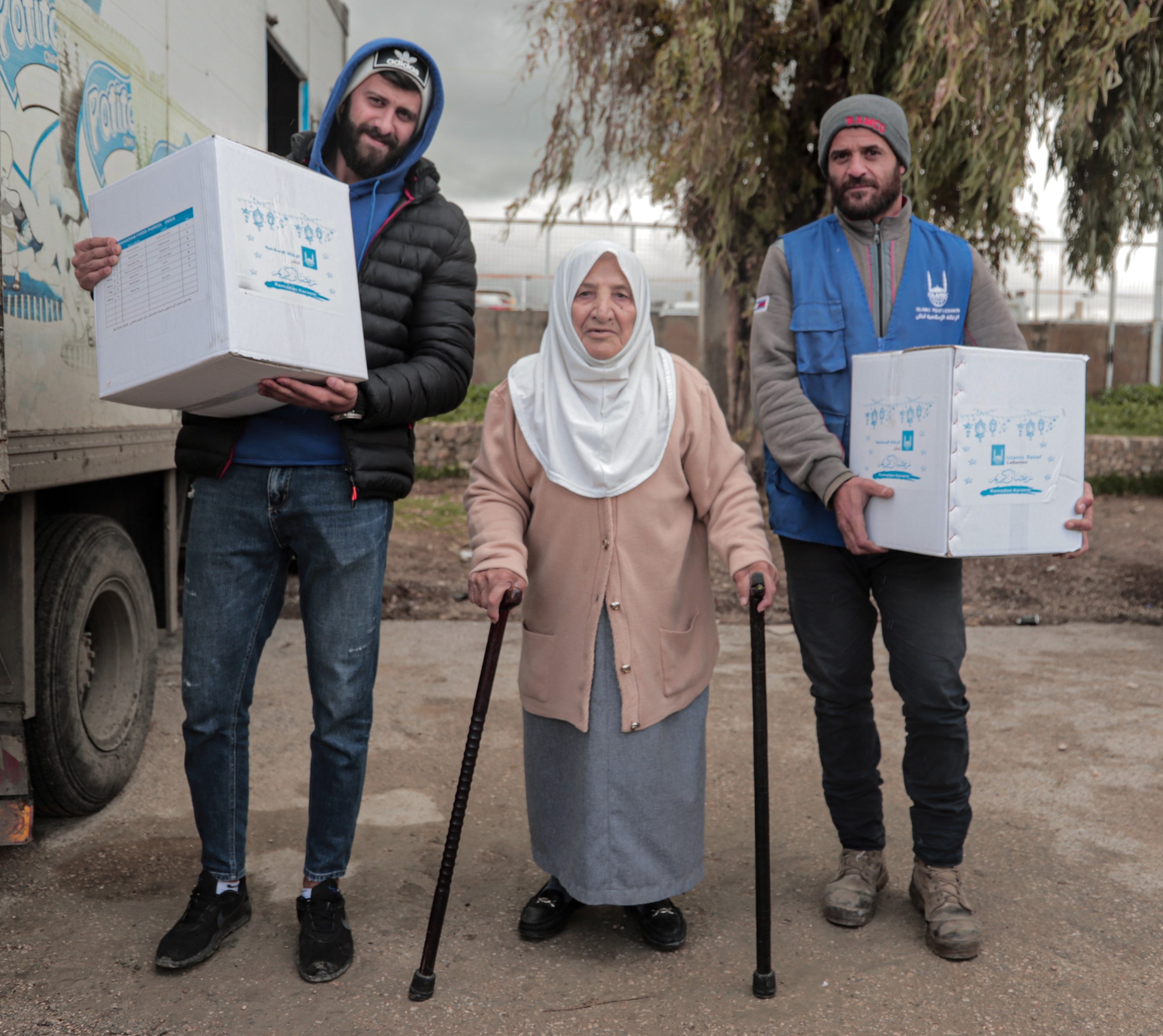 An elderly woman wearing a hijab and leaning on two walking sticks stands next to 2 men carrying food packages.
