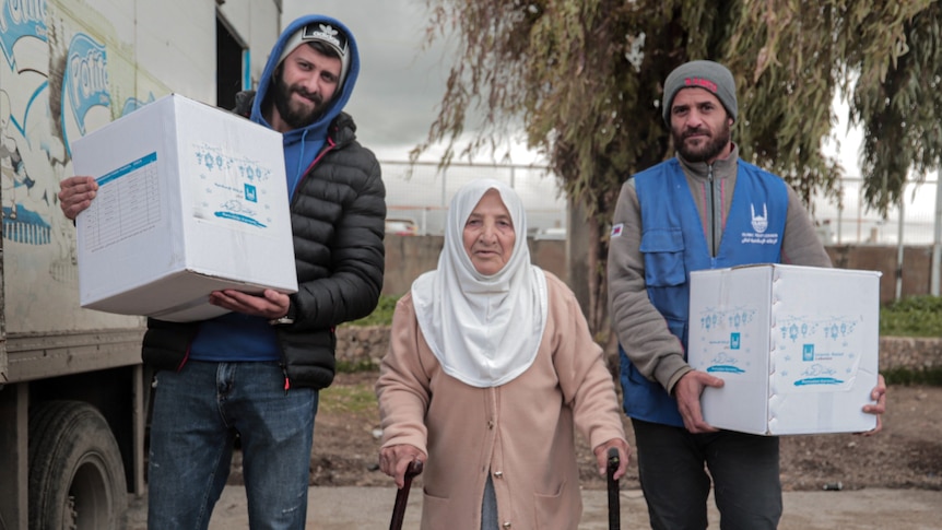 An elderly woman wearing a hijab and leaning on two walking sticks stands next to 2 men carrying food packages.