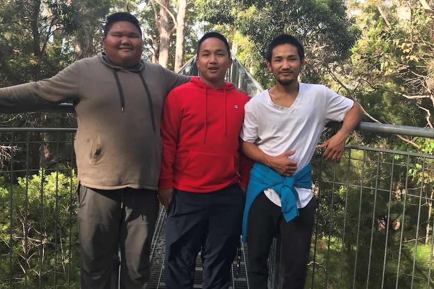 Three Asian teenagers stand together on a bridge with rainforest behind them