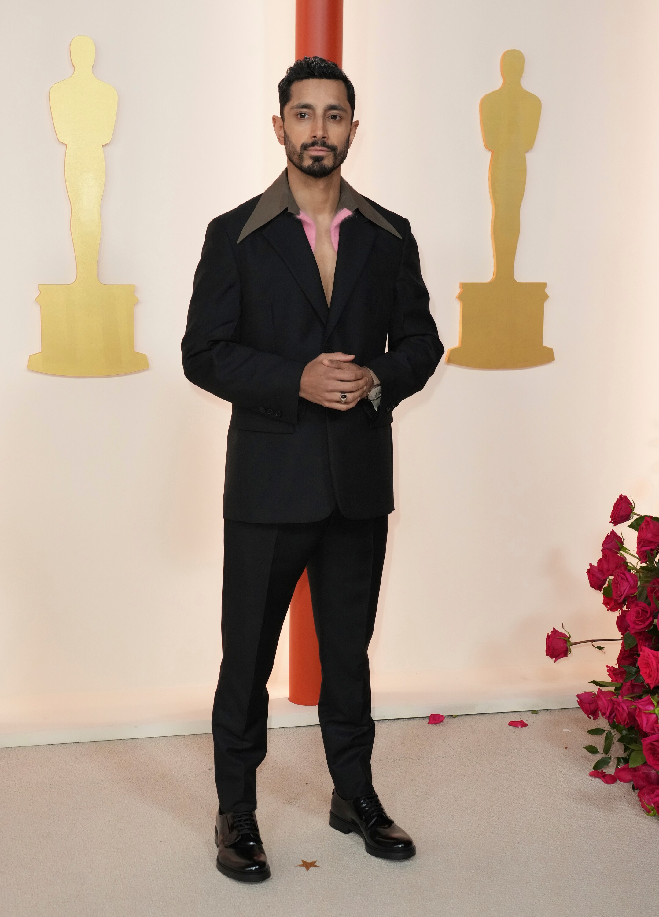 Riz Ahmed wearing a black suit jacket with a pale pink collar detailing and black trousers.
