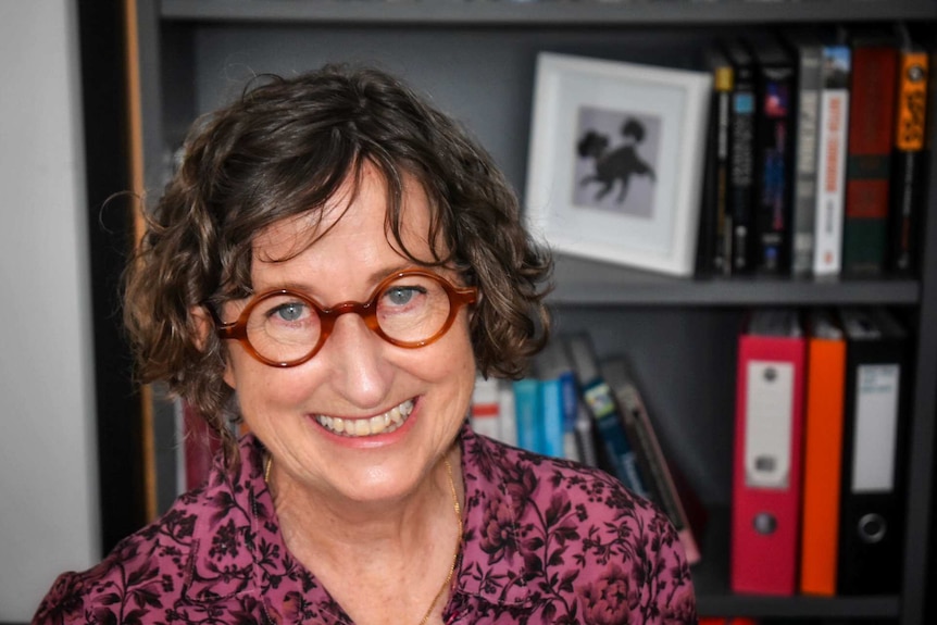 Middle aged woman who has short, dark curly hair and round glasses, with smile.