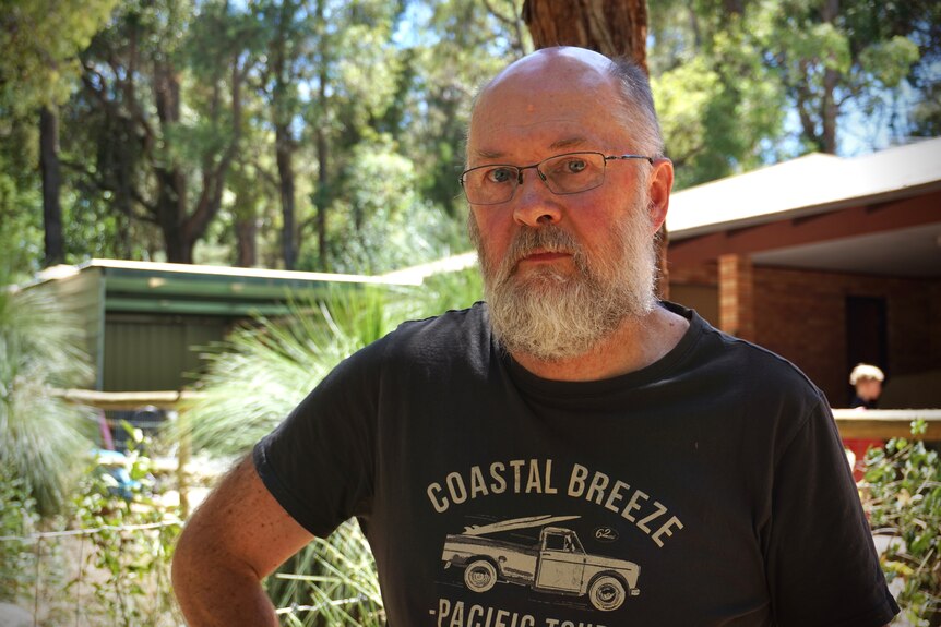 A man with a stern expression, white beard and glasses stands in front of a home in the Perth hills