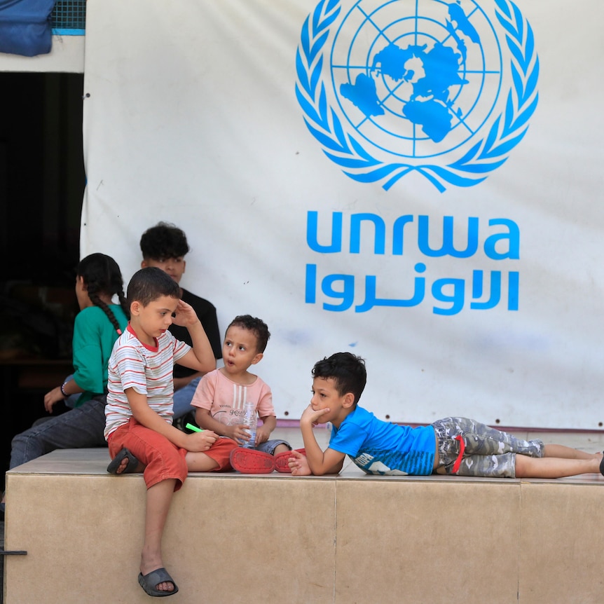 A group of young children lounge in front of a blue and white banner for UNRWA.