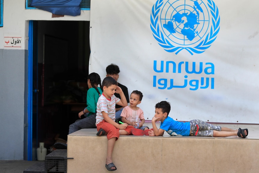 A group of young children lounge in front of a blue and white banner for UNRWA.