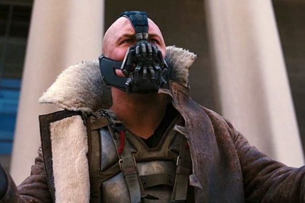 Bane spreads his hands wide as he stands in front of the pillars of a building.