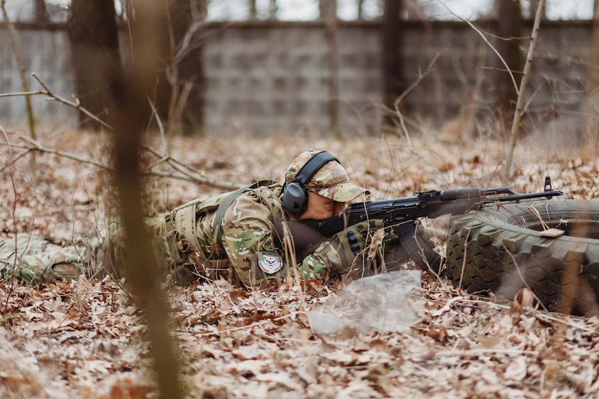 A man in military gear lays on brown leaves, leaning his rifle on an old tyre, lining up a shot