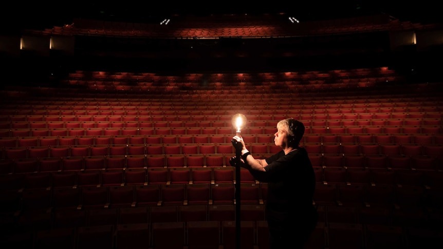 A woman adjusts the light bulb of a lamp standing on stage. A large, empty auditorium of red seats can be seen behind her.