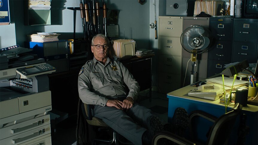 An older man wears glasses and law enforcement uniform sits slumped in chair in dimly lit office.