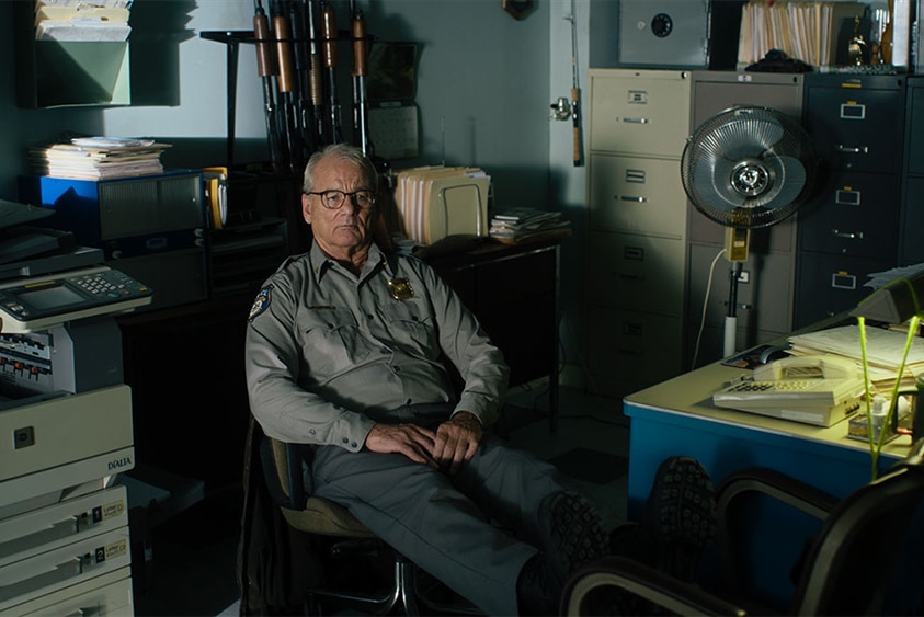 An older man wears glasses and law enforcement uniform sits slumped in chair in dimly lit office.