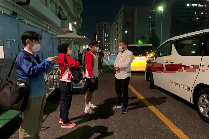 Two athletes in red and white stand on footpath next to man in white with face mask in front of white van at night.