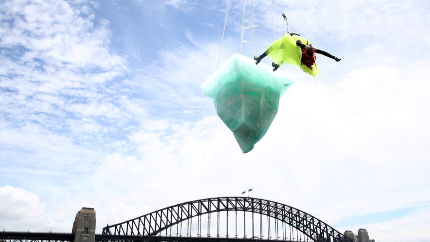 An Aboriginal woman is suspended with her heeled boots on the edge of a melting iceberg, which hangs over Sydney Harbour