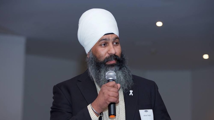 An older man, wearing a turban and sporting a long a grey and black beard, speaks into a microphone.