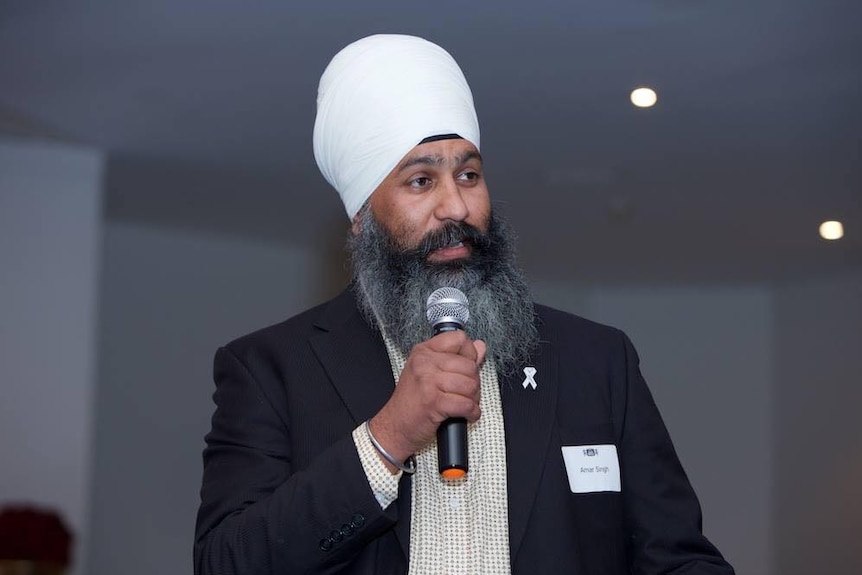 An older man, wearing a turban and sporting a long a grey and black beard, speaks into a microphone.