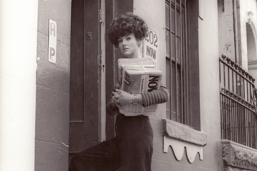 A black and white photo of a woman in a striped top and black pants holding a newspaper titled NOW.