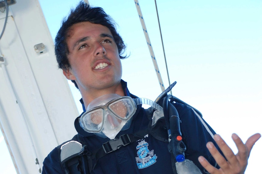 A mid-shot on an angle of Jarrod Hampton wearing diving goggles around his neck and other diving gear standing on a boat.