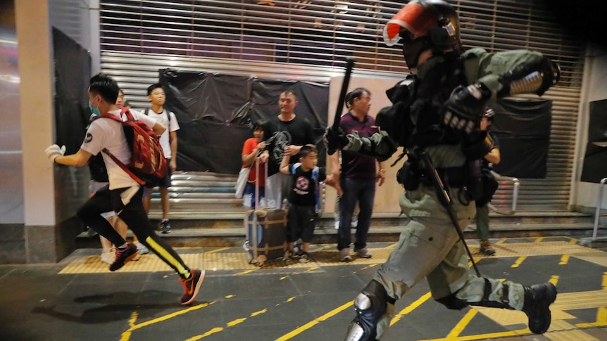 A police officer in riot gear chases protesters through a station as a scared family with children look on