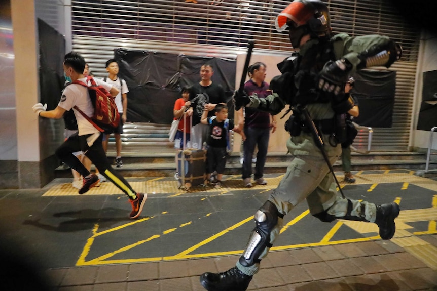 A police officer in riot gear chases protesters through a station as a scared family with children look on