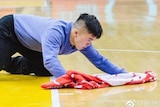 Image of Meng Fei cleaning during a basketball game. Players are standing behind, he is on his hands and knees with a towel.