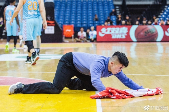 Image of Meng Fei cleaning during a basketball game. Players are standing behind, he is on his hands and knees with a towel.