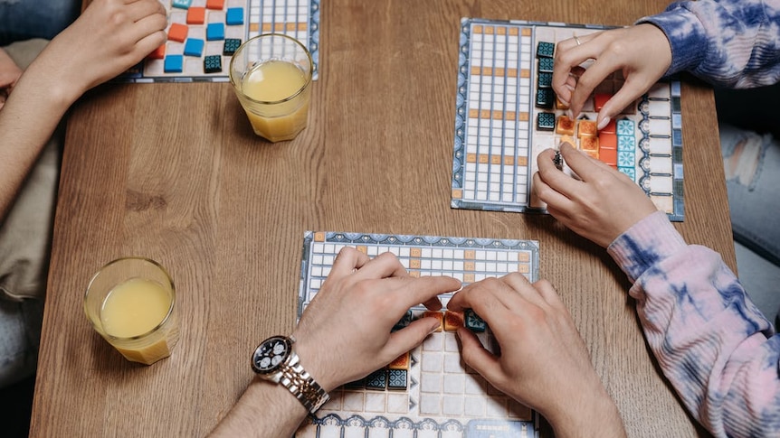 Three people sit around a table with board game pieces in their hands as they play a game. Glasses are on the table with them.