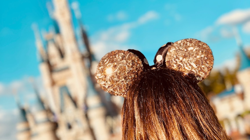 The back of a woman's head wearing glittery gold Mickey Mouse ears as she looks towards a Disney castle.