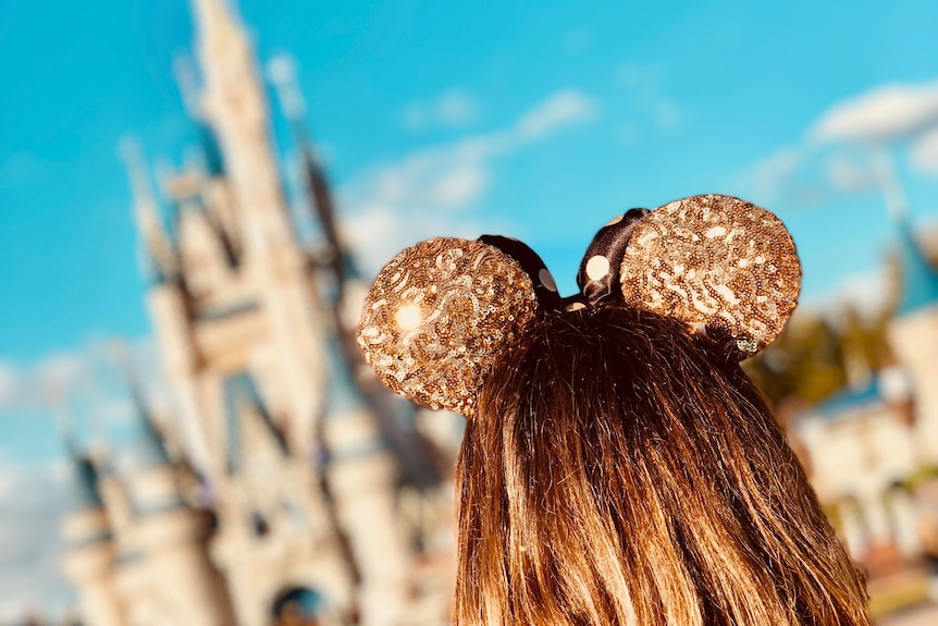 The back of a woman's head wearing glittery gold Mickey Mouse ears as she looks towards a Disney castle.
