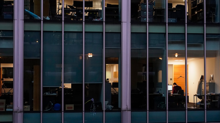 An external shot of a building shows several offices in the dark, with only one window lit up, a worker sitting at a desk inside