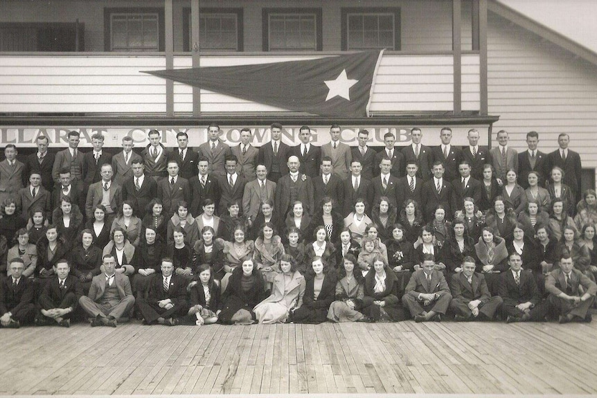 About 100 people pose in ordered rows in front of a rowing club building in the 1930s.
