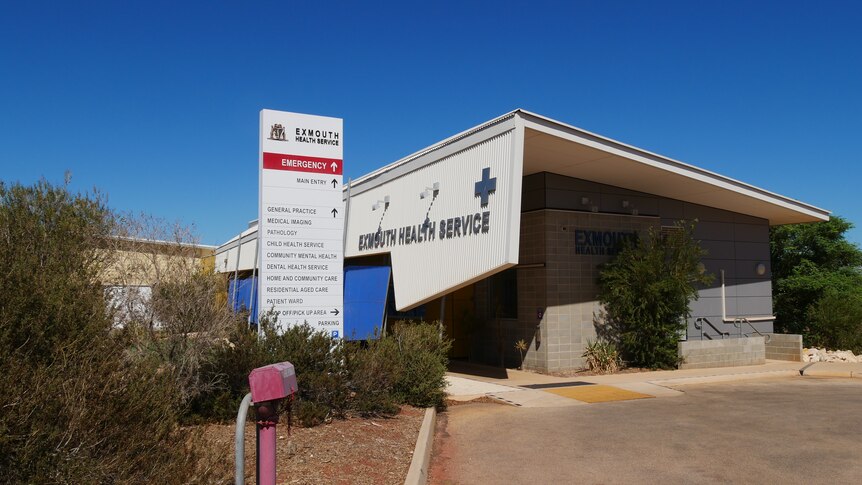 A white sign against blue sky pointing to the hospital's emergency department, in front of a modern corrugated iron building.