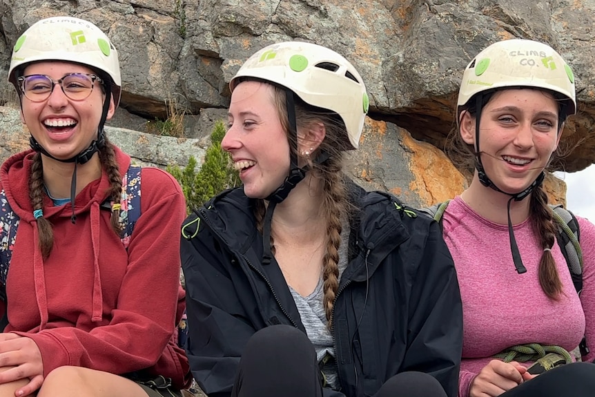 3 girls wearing braids with white helmets sitting on a mountain smiling.