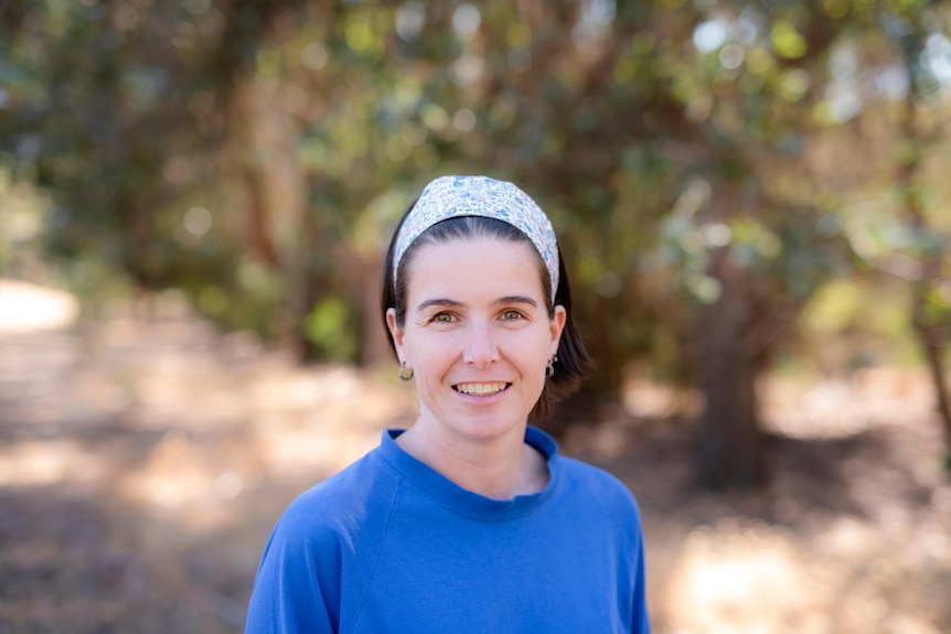 A woman wearing a white headband and blue top outside surrounded by trees.