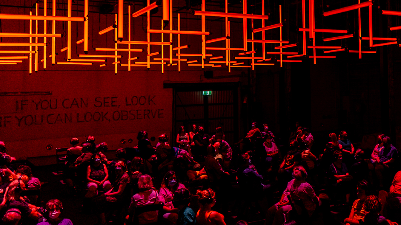 An audience is individually seated in the dark, wearing headphones and face masks, under  a grid of red lights.