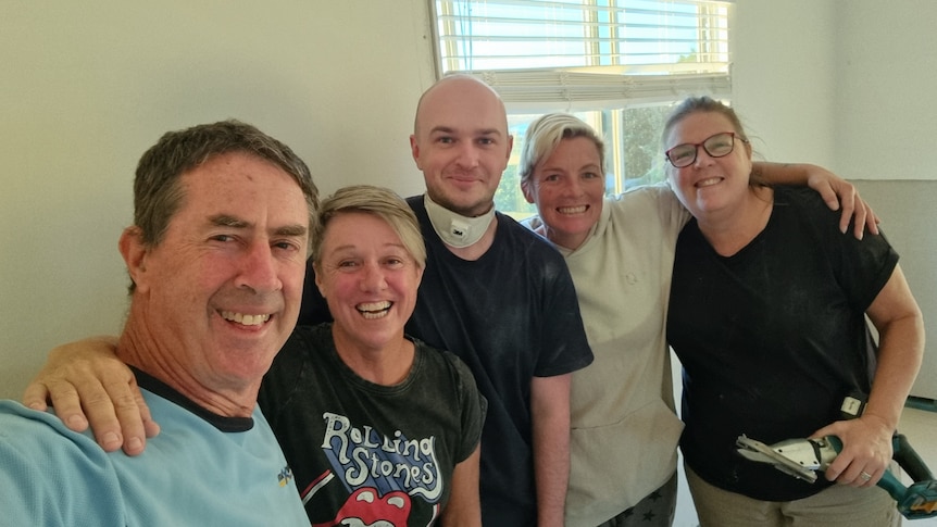 Five people standing together in a white room smiling. Three women, two men. 