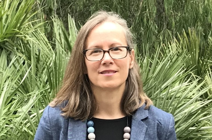 A woman wearing glasses stands in front of green plants and trees looking at the camera on a bright day.