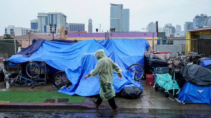 A person wearing a rain poncho walks past things covered in tarpaulin