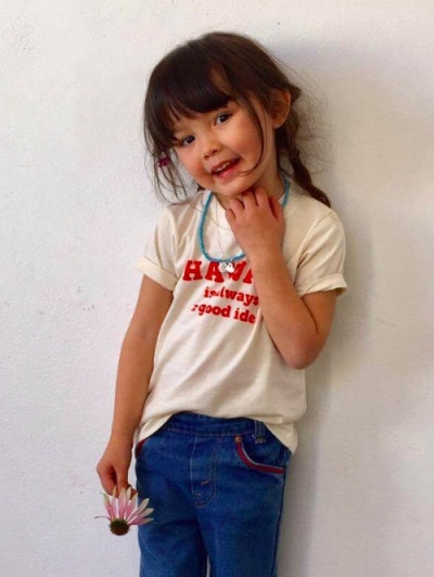 Kawa, wearing jeans, a t-shirt and blue necklace, smiles and holds a flower.