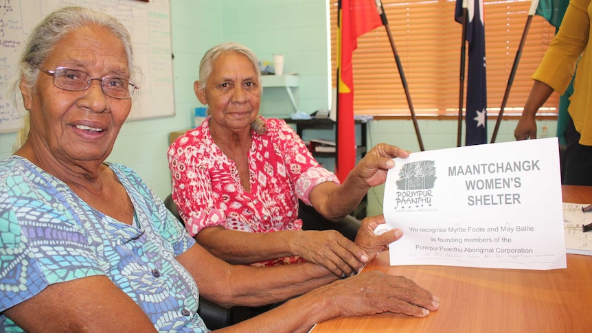 Two older Aboriginal women hold up a piece of paper with 'Pormpur Paanthu Maantchanngk Women's Shelter' written on it.