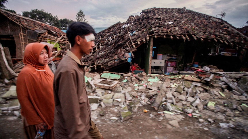 A man and a woman walk past a street littered with rubble and a destroyed house.