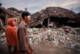 A man and a woman walk past a street littered with rubble and a destroyed house.