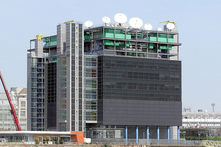 Looking across a brown river stands a black square tower with a multitude of white satellites on its rooftop.