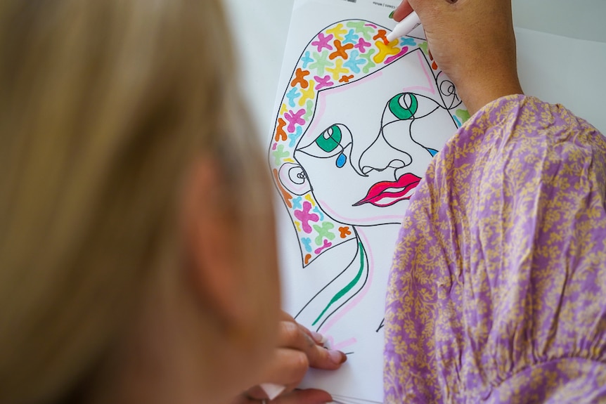 A hand draws a colourful female face with texta.