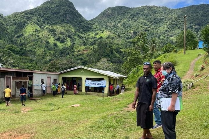 People stand on a hill outside a polling venue in a remote part of fiji with mountains in the background