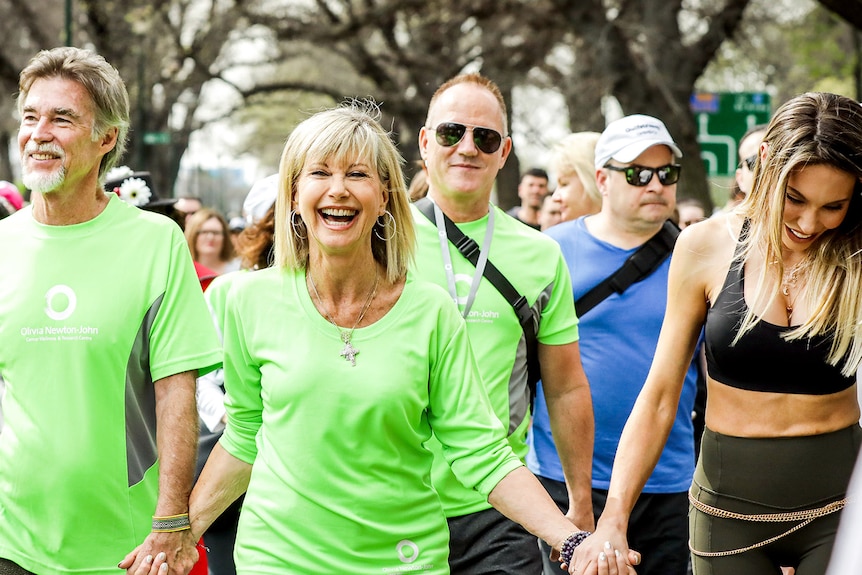 Olivia Newton-John smiling warmly, walking among a crowd of runners.