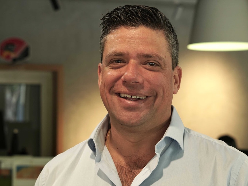 A man with dark hair, wearing a light blue shirt smiles to the camera.