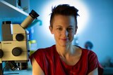 Dr Linda Iles, who has short brown hair and is wearing a red top, poses for a photo next to a microscope in a blue-lit room