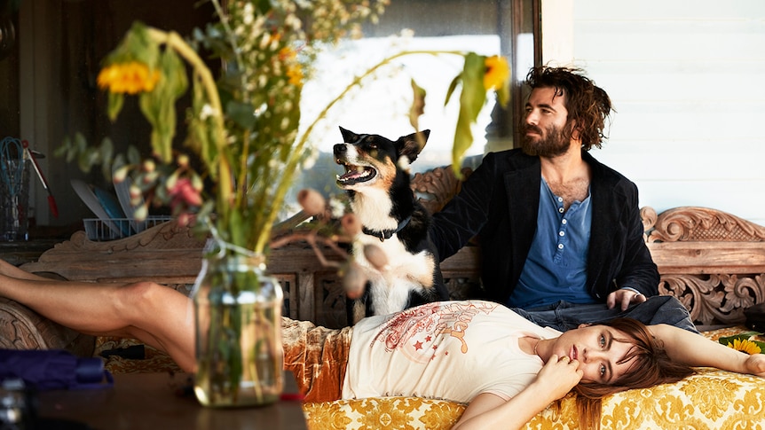 Angus Stone sits on a couch patting a dog, while Julia Stone lies down in front of him